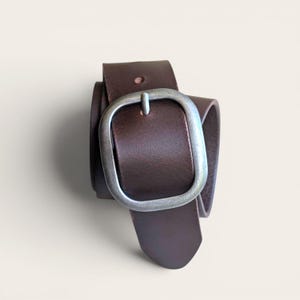 Peut inclure: Une ceinture en cuir marron fonc&eacute; dot&eacute;e d'une grande boucle rectangulaire arrondie avec une finition argent&eacute;e mate. La sangle de la ceinture est lisse et de couleur unie, avec un seul trou de r&eacute;glage visible pr&egrave;s de la boucle.