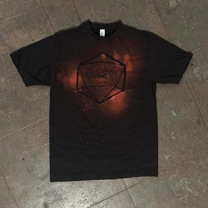 May include: Black t-shirt featuring a geometric design of a 20-sided die with the word "DIE" inside. The design has a red and orange spray-paint effect. The shirt is a crew neck.
