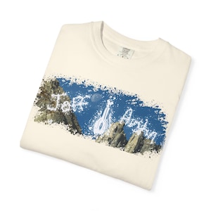 May include: Cream-coloured t-shirt with a blue and white graphic. The design features a guitar, mountains, and the text "Jeff Austin". The shirt is folded.