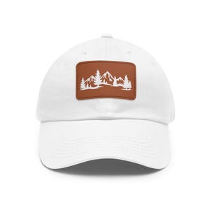May include: A white baseball cap featuring a rectangular brown patch centered on the front. The patch displays a white graphic illustration of a mountain range with several pine trees in the foreground, stitched with visible brown thread.