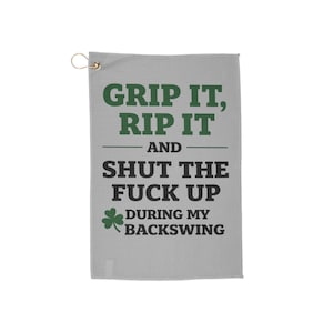 May include: A light gray golf towel with the text "GRIP IT, RIP IT AND SHUT THE FUCK UP DURING MY BACKSWING" in green and black. A gold-colored metal hook is attached to the top left corner. A green shamrock is also featured.