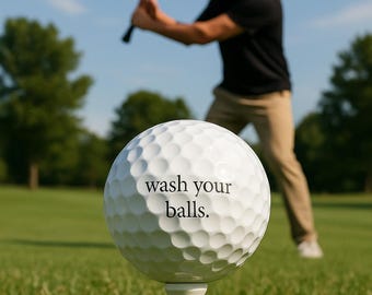 May include: A white golf ball on a tee with the text "wash your balls." A person in a black shirt and khaki pants is swinging a golf club in the background on a green golf course.