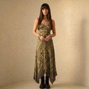 May include: A sleeveless midi dress featuring an olive green and dark brown paisley bandana print with a handkerchief hemline. The dress has a scoop neckline and is paired with black lace-up combat boots, layered silver necklaces, and multiple rings.