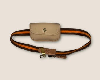 May include: A tan leather fanny pack with a brown and orange striped belt. The belt has a gold-coloured buckle and a small leather strap. The fanny pack has a snap closure.