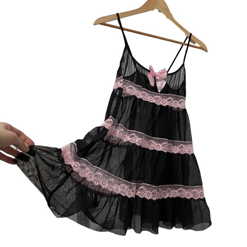 May include: Black babydoll lingerie with spaghetti straps and a pink bow. The sheer nightgown features pink lace trim and a tiered design. The garment is hanging on a wooden hanger.