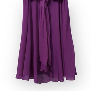 May include: A floor-length, flowing purple skirt made of a lightweight fabric. The skirt features a high waist with a tie detail and a flared silhouette. The fabric drapes gracefully, creating soft folds and movement.