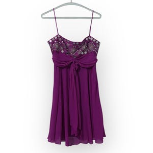 May include: A sleeveless, knee-length purple dress with thin spaghetti straps. The bodice is embellished with silver sequins and beads. A large bow detail sits at the waist, and the skirt flows down from the bow. The dress hangs on a gray hanger.