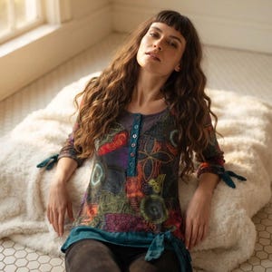 Puede incluir: Una blusa de manga larga con base en color verde azulado oscuro y un patrón abstracto multicolor en tonos rojo, naranja, verde y morado. La prenda incluye una tapeta con botones en el cuello y detalles de lazos en las mangas y el dobladillo.