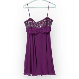 May include: A purple dress with thin spaghetti straps, hanging on a light green hanger. The bodice is embellished with silver sequins and beads. The dress has a gathered waist and a flowing skirt, perfect for a special occasion.