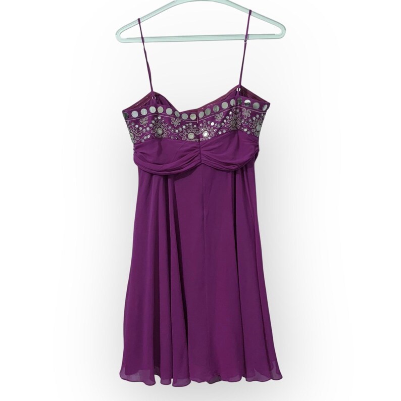 May include: A purple dress with thin spaghetti straps, hanging on a light green hanger. The bodice is embellished with silver sequins and beads. The dress has a gathered waist and a flowing skirt, perfect for a special occasion.