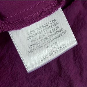 May include: Close-up of a white clothing tag with black text on a deep purple fabric. The tag lists fabric composition: 100% silk and 100% polyester. The tag also includes the text "CARE ON REVERSE".