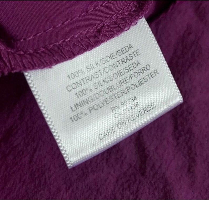 May include: Close-up of a white clothing tag with black text on a deep purple fabric. The tag lists fabric composition: 100% silk and 100% polyester. The tag also includes the text "CARE ON REVERSE".