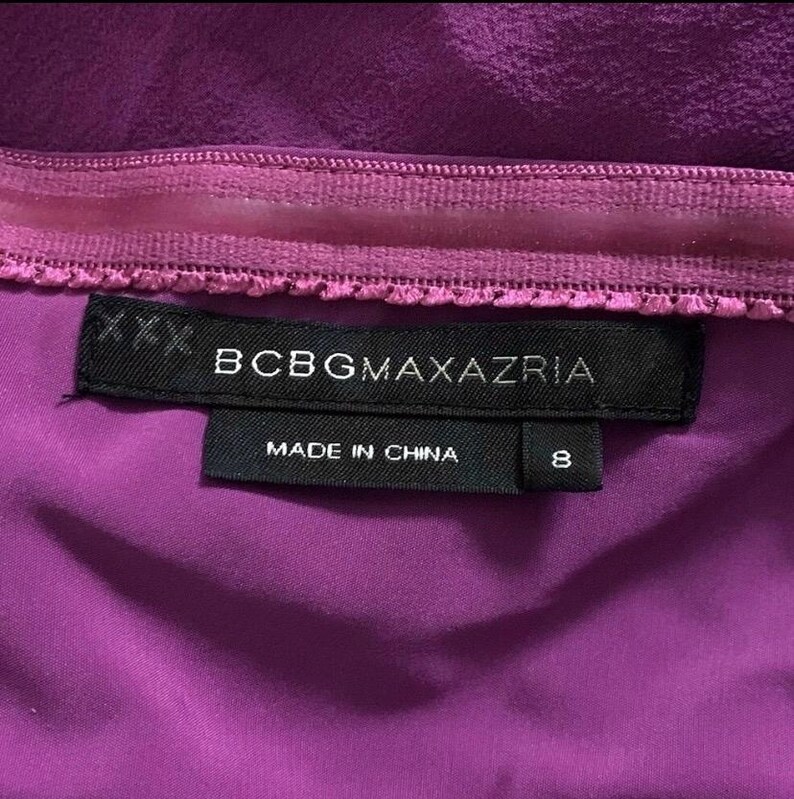 May include: Close-up of a purple garment with a stitched seam and a black label. The label reads "BCBGMAXAZRIA" and "MADE IN CHINA" with a size 8. The fabric is a solid, deep purple color.