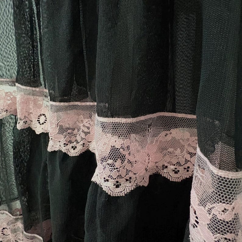 May include: Close-up of black sheer fabric with delicate pink lace trim. The lace features a floral design, adding a touch of elegance. The fabric is layered, creating depth and texture. The overall aesthetic is vintage and romantic.
