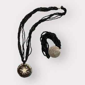 Puede incluir: Un conjunto de collar y pulsera de cuentas negras con un gran colgante redondo que presenta un diseño de estrella de mar tallada. El colgante está hecho de nácar y tiene un fondo marrón.