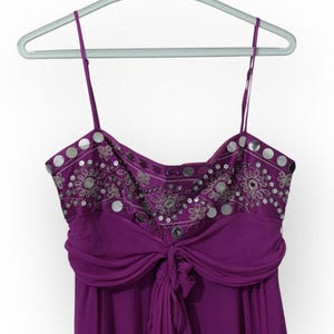 May include: A purple formal dress with thin straps, hanging on a hanger. The bodice features silver sequins and embroidery. The dress has a draped waist and a flowing skirt, perfect for special occasions.