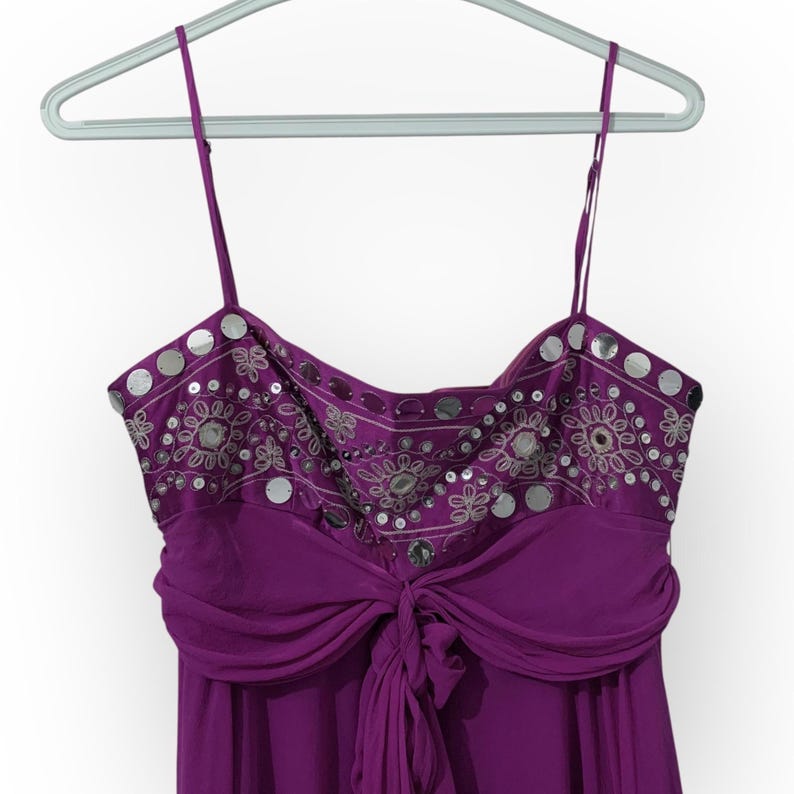 May include: A purple formal dress with thin straps, hanging on a hanger. The bodice features silver sequins and embroidery. The dress has a draped waist and a flowing skirt, perfect for special occasions.