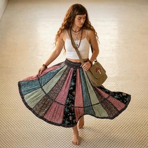 May include: A white sleeveless crop top paired with a multi-coloured patchwork skirt featuring vertical panels of floral and patterned fabric in shades of pink, blue, green, and black. A tan crossbody bag with a small mushroom patch is worn.