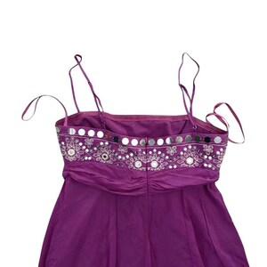 May include: A sleeveless, purple dress with thin straps. The bodice features a decorative band with silver sequins and embroidered floral patterns. The dress has a gathered waist and a flowing skirt.