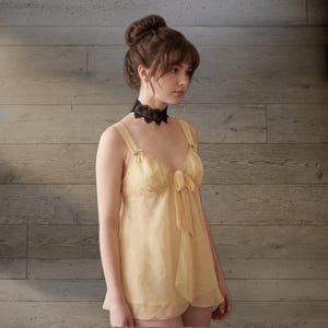May include: A light yellow camisole with spaghetti straps and a bow detail. The camisole is paired with a black lace choker. The background is a wooden wall.