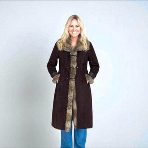 May include: A dark brown coat with faux fur trim on the collar, cuffs, and down the front. The coat has a long, tailored silhouette and is worn with blue jeans. The woman is smiling.