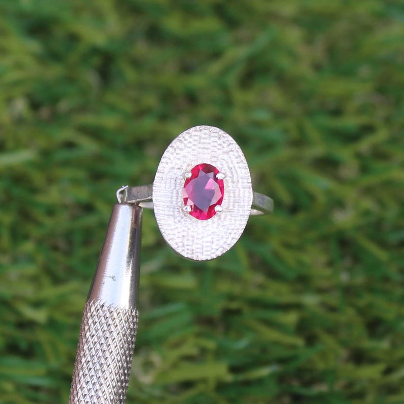 May include: A silver ring with an oval, textured setting. The setting surrounds a vibrant, oval-cut, red gemstone. The ring is held by a tool against a blurred green background, showcasing the jewelry's design.