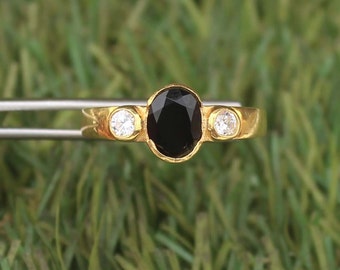May include: A gold ring featuring a large, oval-cut black gemstone in the center, flanked by two smaller, round, clear gemstones. The ring is held by tweezers against a green background.