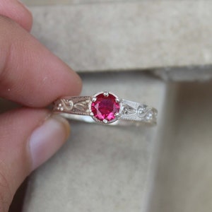 May include: A silver ring with a round, bright red gemstone. The band features an engraved floral pattern. The ring is held up, showcasing the details of the design and the vibrant color of the stone.