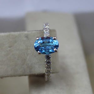 Op de afbeelding: Een zilveren ring met een grote, ovaalvormige, helderblauwe edelsteen. De band is bezet met kleine, heldere edelstenen. Het ontwerp van de ring benadrukt het levendige blauw van de centrale steen tegen een neutrale achtergrond.