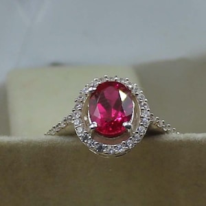 Puede incluir: Anillo de plata con una gran gema de rubí de corte ovalado. El rubí está rodeado por un halo de piedras preciosas más pequeñas y transparentes. La banda del anillo también está adornada con pequeñas piedras preciosas transparentes, creando un efecto brillante.