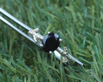May include: A silver ring with a large, oval-cut black gemstone. The ring's band features a textured, branch-like design. The ring is set against a backdrop of green grass.