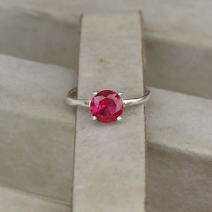 Op de afbeelding: Een zilveren ring met een grote, ronde, helderrode edelsteen. De edelsteen is het middelpunt, gezet in een eenvoudig, klassiek ontwerp. De ring wordt getoond tegen een neutrale achtergrond, waardoor de kleur en glans van de edelsteen worden benadrukt.