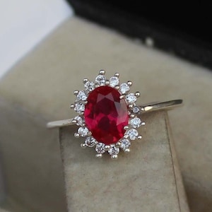 Op de afbeelding: Zilveren ring met een grote, ovaal geslepen robijn, omringd door een halo van kleinere, heldere edelstenen. De robijn is helderrood en de ring wordt gepresenteerd tegen een neutrale achtergrond.