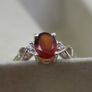 May include: A silver ring with an oval, reddish-brown gemstone at its centre. Two small, clear gemstones are either side of the main stone. The ring's band has a decorative, swirling design.