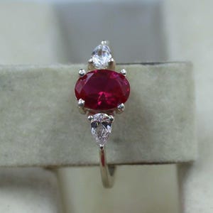Peut inclure: Une bague en argent ornée d'une grande pierre rouge foncé de taille ovale en son centre. Deux petites pierres claires, en forme de poire et rondes, flanquent la pierre principale. L'anneau de la bague est fin et poli.
