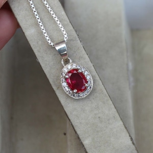 Pode incluir: Um colar com pingente de prata com uma vibrante pedra preciosa vermelha oval, rodeada por pequenas pedras transparentes. O pingente pende de uma delicada corrente de prata. O colar é exibido em uma superfície de tom neutro.