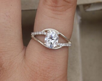 Peut inclure: Une bague en argent ornée d'une grosse pierre précieuse transparente de forme ovale en son centre. La monture se divise en deux bras délicats qui entourent la pierre principale, avec de plus petites pierres transparentes serties dans la monture de chaque côté de la pierre principale.
