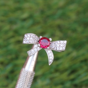 May include: A silver-toned bow ring with a large, round, red gemstone at the center. The bow is encrusted with small, clear gemstones. The ring is held by a silver tool against a blurred green background.