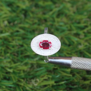 May include: A silver ring featuring an oval, deep red gemstone set in an oval textured setting. The ring is displayed against a green background, showcasing the jewelry's design and color.