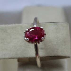 May include: A silver ring with a large, oval ruby gemstone. The ruby is a deep red hue, set in a crown-style setting. The ring's band is slender and polished, showcasing the gemstone.