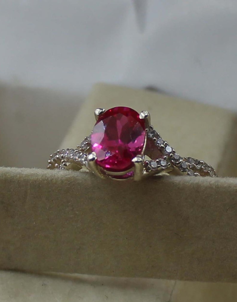 May include: A gold-coloured ring with a large, oval-cut, bright pink gemstone as the focal point. The band is embellished with smaller, clear gemstones, adding sparkle. The ring is set against a neutral background, highlighting its design.