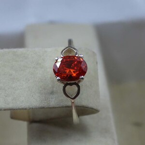 May include: A silver ring featuring a large, oval-cut, fiery orange gemstone. The ring's band has a delicate design with heart-shaped cutouts near the gemstone and at the top of the band, adding a touch of romance to the jewelry.