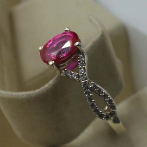 May include: A gold ring with a large, oval-cut, bright pink gemstone. The band has a twisted design, embellished with small, clear accent stones. The ring is set against a neutral background. The design creates a sparkling effect.