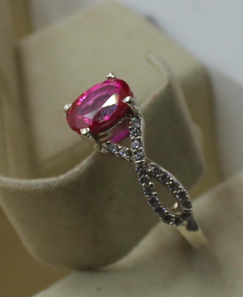 May include: A gold ring with a large, oval-cut, bright pink gemstone. The band has a twisted design, embellished with small, clear accent stones. The ring is set against a neutral background. The design creates a sparkling effect.