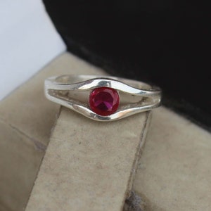 May include: A silver ring with a bright red gemstone. The ring has a split band design that curves around the gemstone, creating a distinctive and elegant appearance. The round gemstone is securely set within the ring's design, adding a pop of colour.
