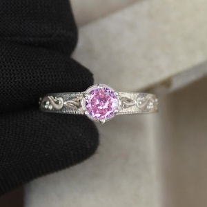 May include: A silver ring with a round, light pink gemstone. The band has an ornate, engraved design. The ring is set against a neutral background, highlighting the intricate details of the jewellery.