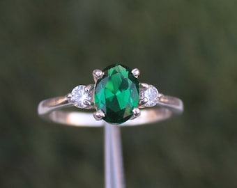 May include: A silver ring featuring a large, oval-cut emerald green gemstone as the centerpiece, flanked by two smaller, round, clear gemstones. The ring is set against a blurred green background, highlighting the jewelry's sparkle and design.