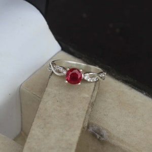 Könnte beinhalten: Ein silberner Ring mit einem leuchtend roten Edelstein in der Mitte. Das Band hat ein verdrehtes Design mit kleinen, klaren Akzentsteinen. Der Ring wird auf einer neutral getönten Oberfläche präsentiert.
