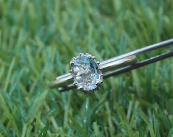 May include: A silver ring with an oval, light blue gemstone. The gemstone is set in a decorative, silver bezel. The ring is held by a pair of silver tweezers against a backdrop of green grass.