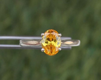 May include: A silver ring featuring a vibrant, oval-cut yellow gemstone. The gemstone is held in place by four prongs. The ring is set against a blurred green background.